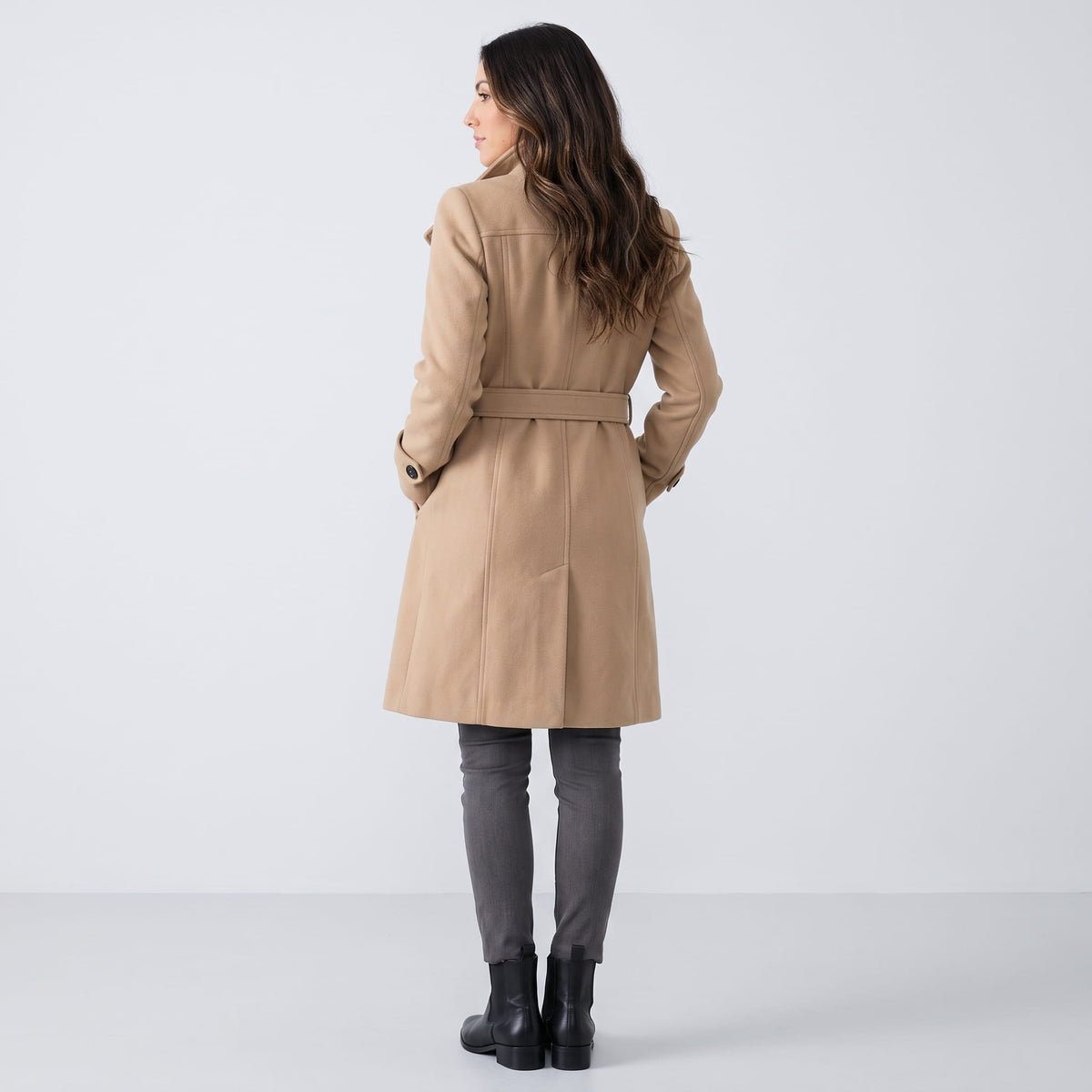 A woman wearing a long beige trench coat with a belted waist and double-breasted front, shown from the back