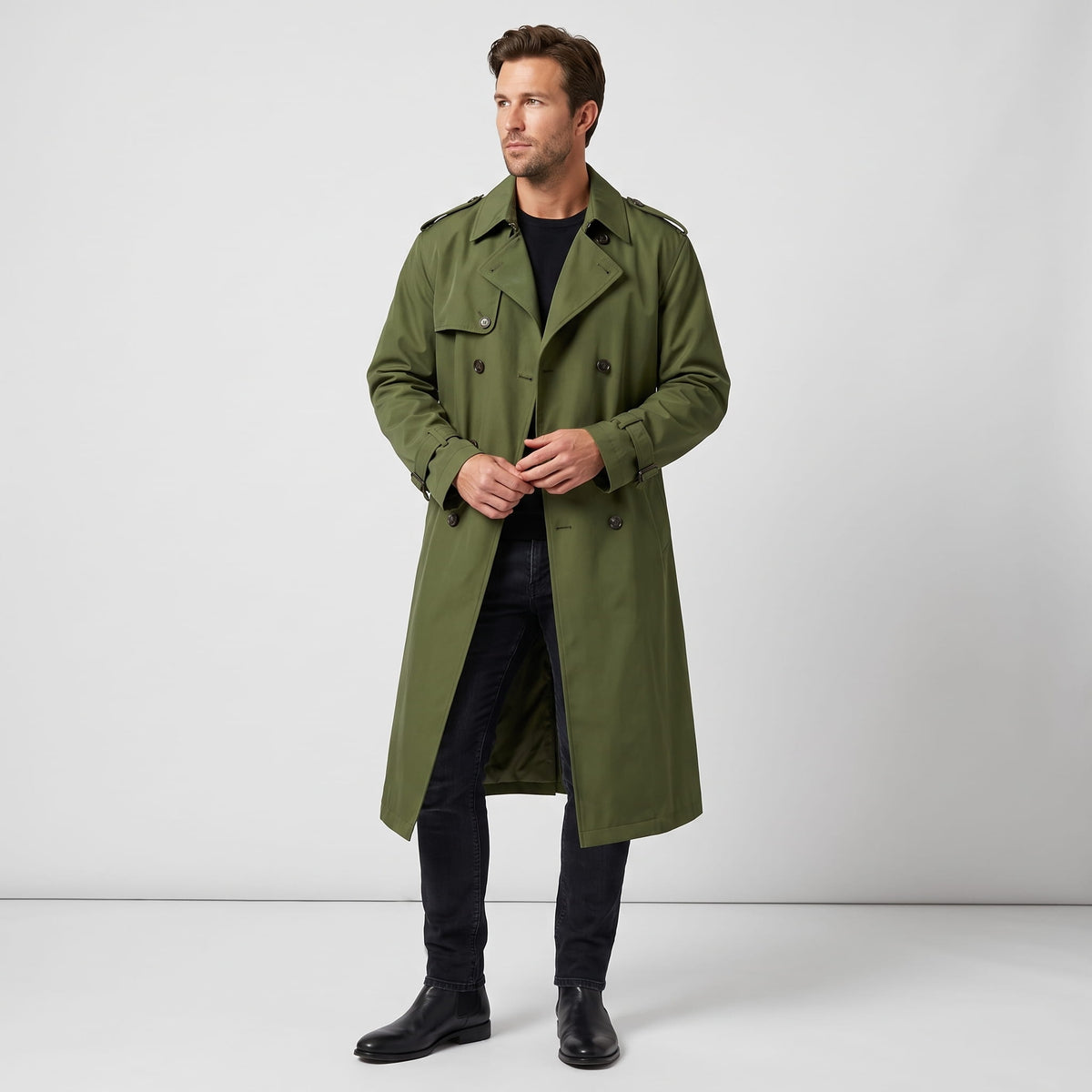 A man wearing a long olive green trench coat with a double-breasted front and waist belt, front view