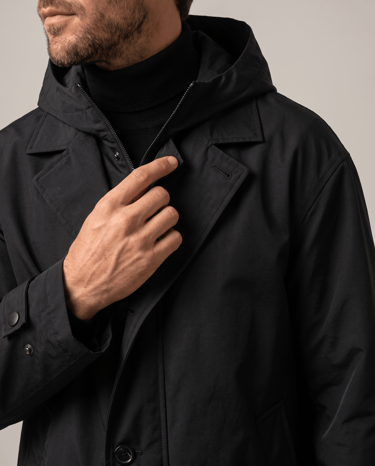Close-up of a man wearing a black longline trench coat, showing the hood, collar, and front closure