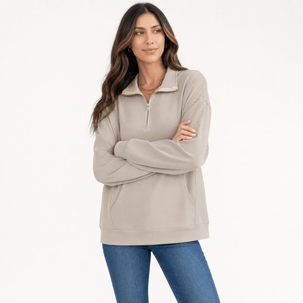 Female model wearing a light taupe quarter-zip sweater with a relaxed fit, paired with blue jeans, arms crossed.
