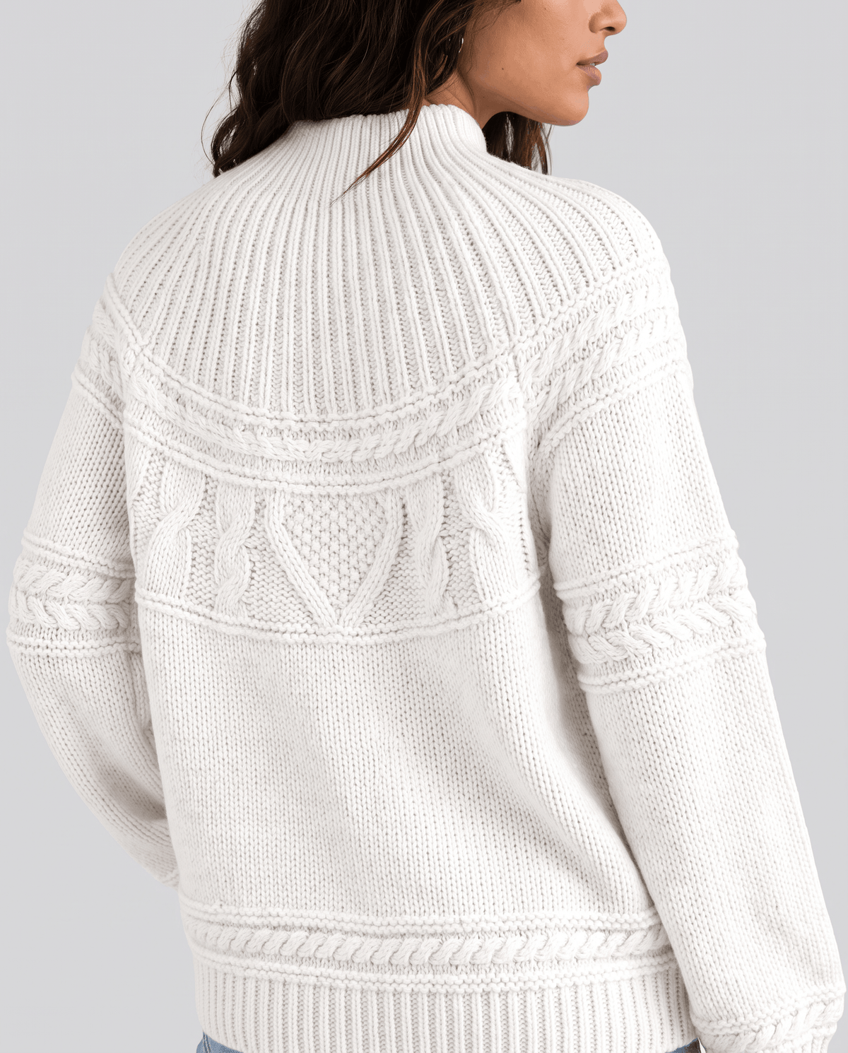 White Merino Wool Cable Knit Sweater worn by a female model from the back, featuring a relaxed fit, textured cable knit pattern, high neckline, and ribbed trim.