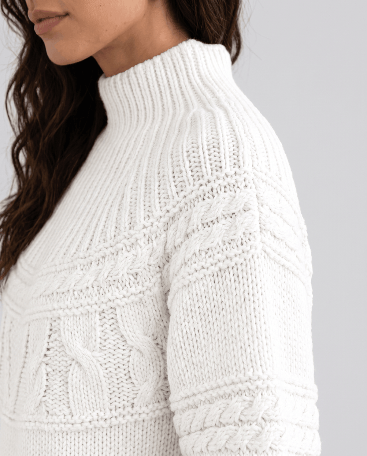 White Merino Wool Cable Knit Sweater worn by a female model, shown in close-up from the side to highlight the cable knit texture, high neckline, and relaxed fit.