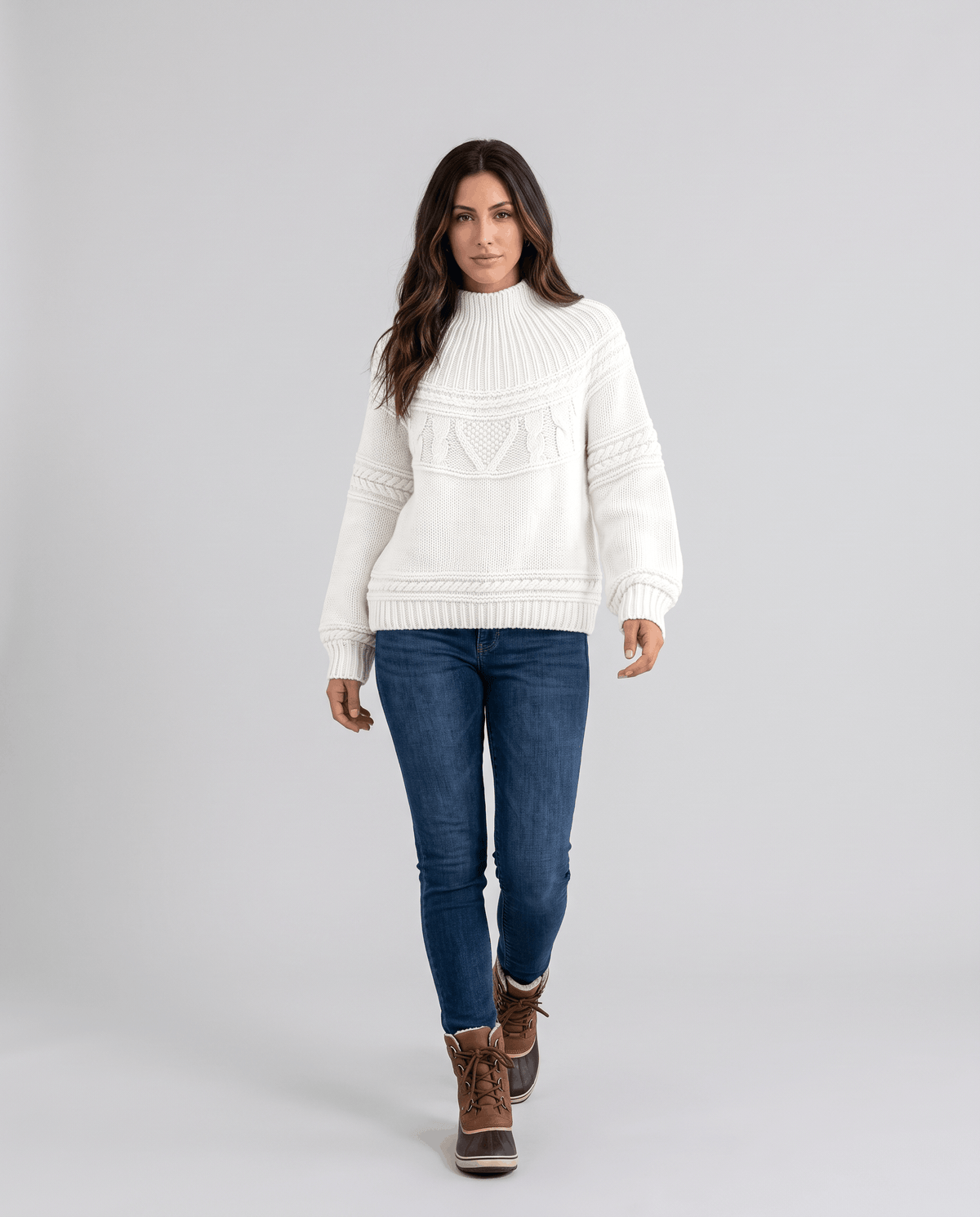 White Merino Wool Cable Knit Sweater worn by a female model with blue jeans, featuring a relaxed fit, textured cable knit pattern, high neckline, long sleeves, and ribbed trim.