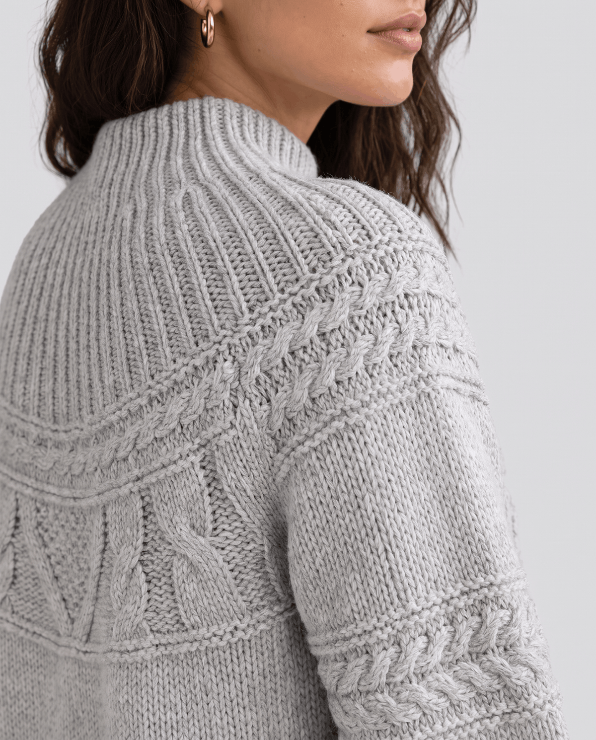 Gray Merino Wool Cable Knit Sweater worn by a female model, shown from the side to highlight the textured cable knit pattern, relaxed fit, and ribbed trim.