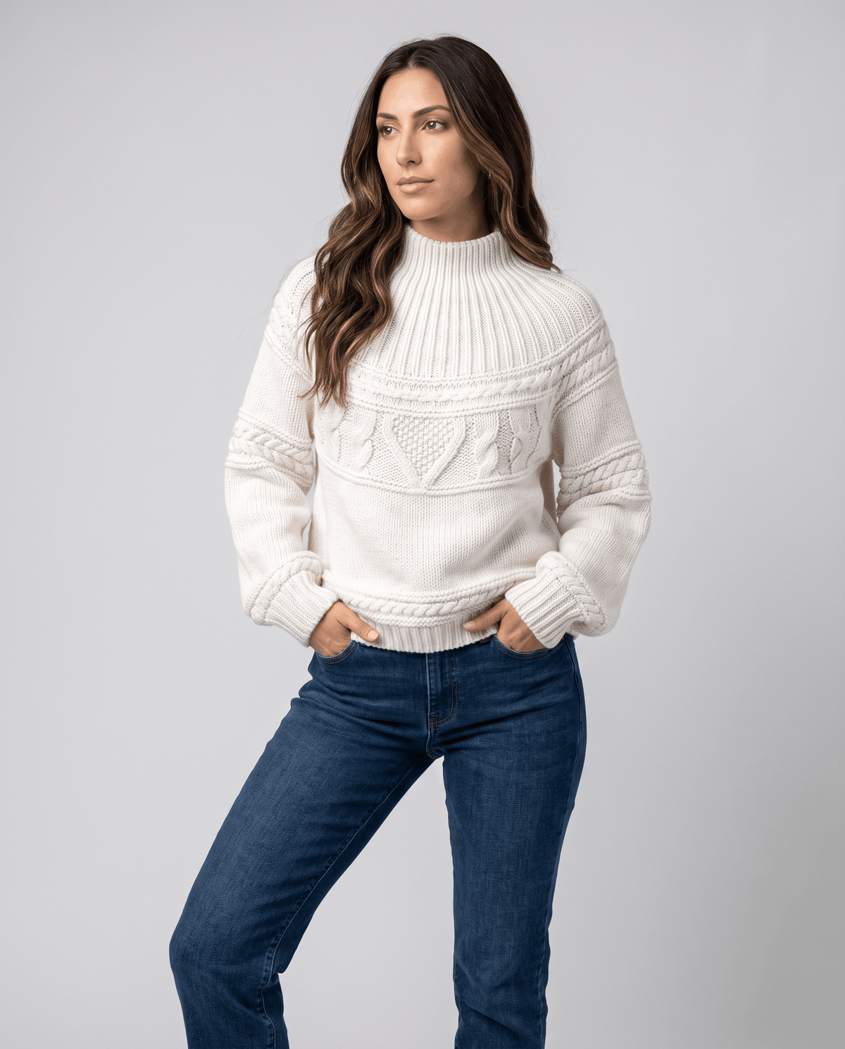 White Merino Wool Cable Knit Sweater worn by a female model with blue jeans, featuring a relaxed fit, textured cable knit pattern, high neckline, and ribbed trim.