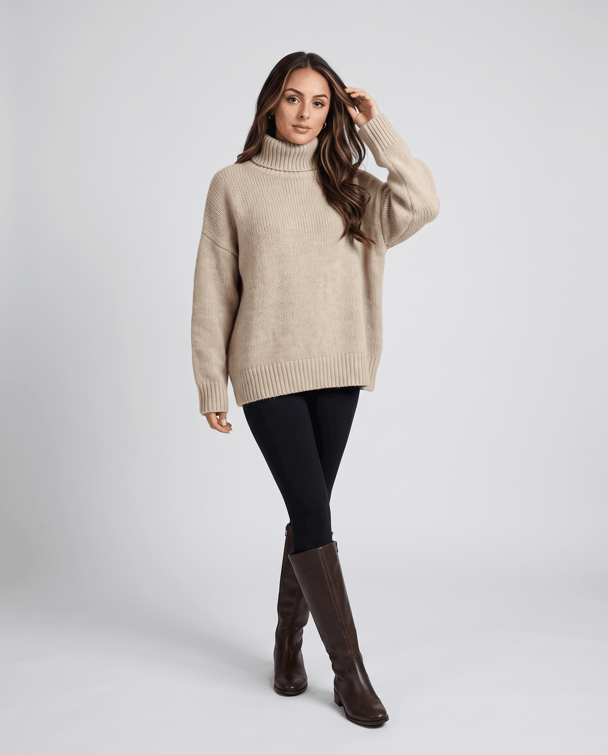Beige Merino Wool Turtleneck Sweater worn by a female model with dark leggings, featuring an oversized fit, high collar, long sleeves, and ribbed cuffs.