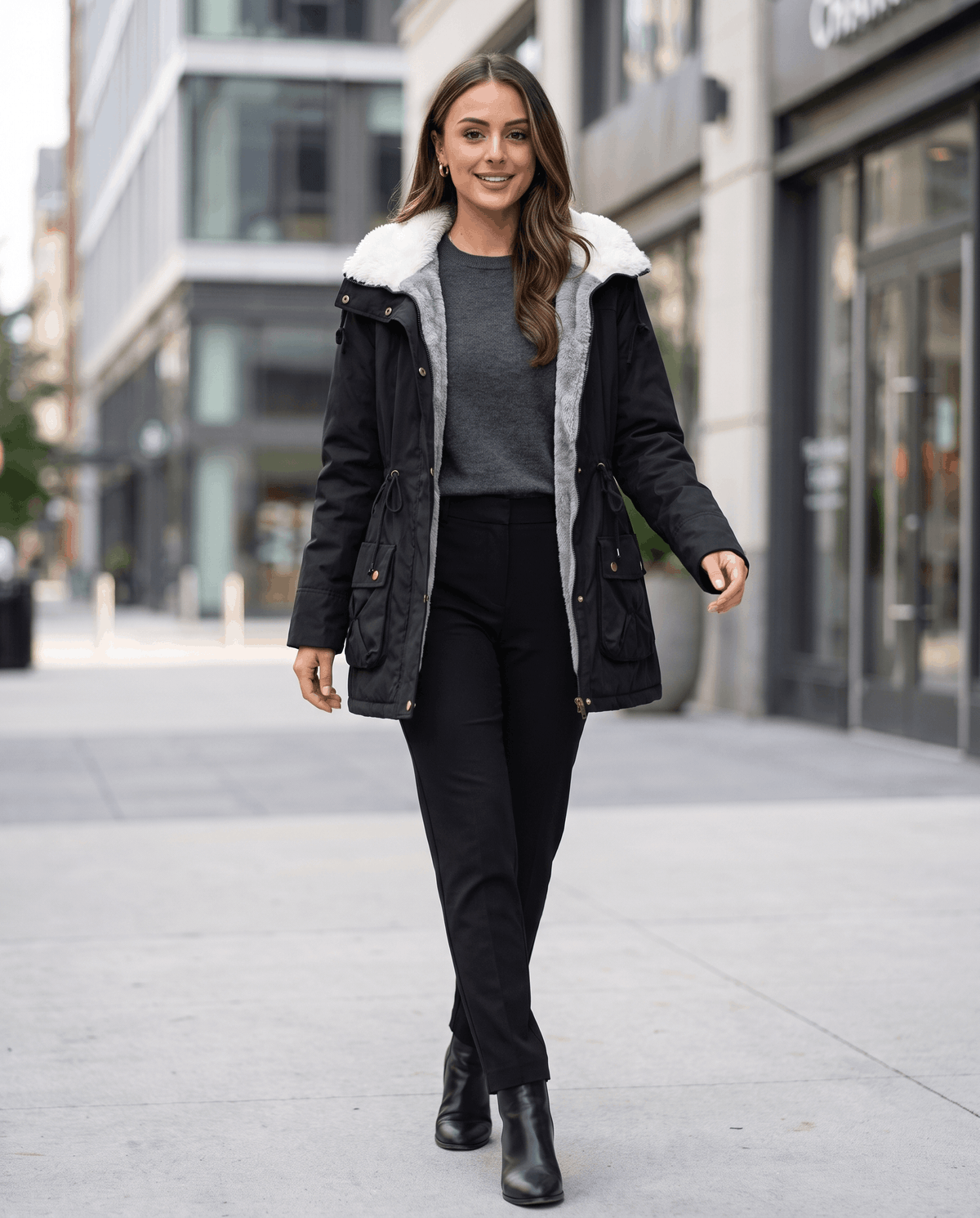 A woman wearing a black mid-length parka jacket with a fur-trimmed hood and drawstring waist, walking on a city street