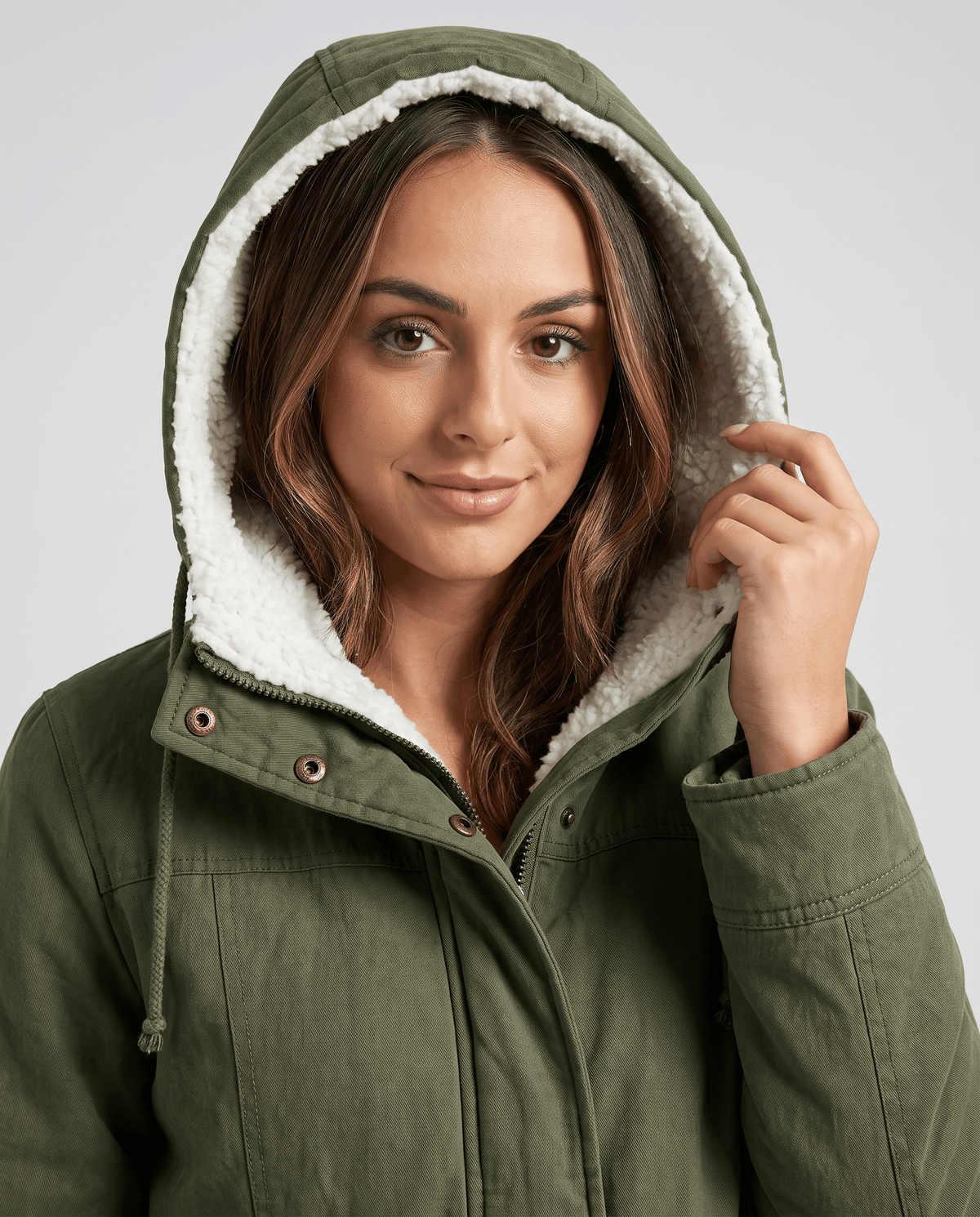 A woman wearing an olive green mid-length parka jacket with a fur-trimmed hood and drawstring waist, close-up view