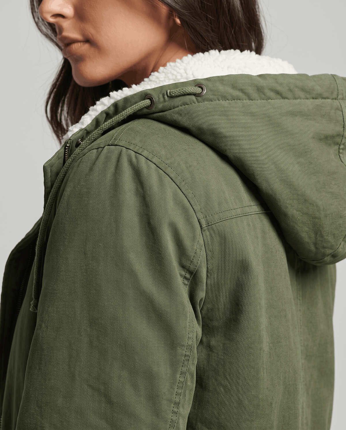 Close-up of a woman wearing an olive green parka jacket, showing the fur-trimmed hood and drawstring waist detail