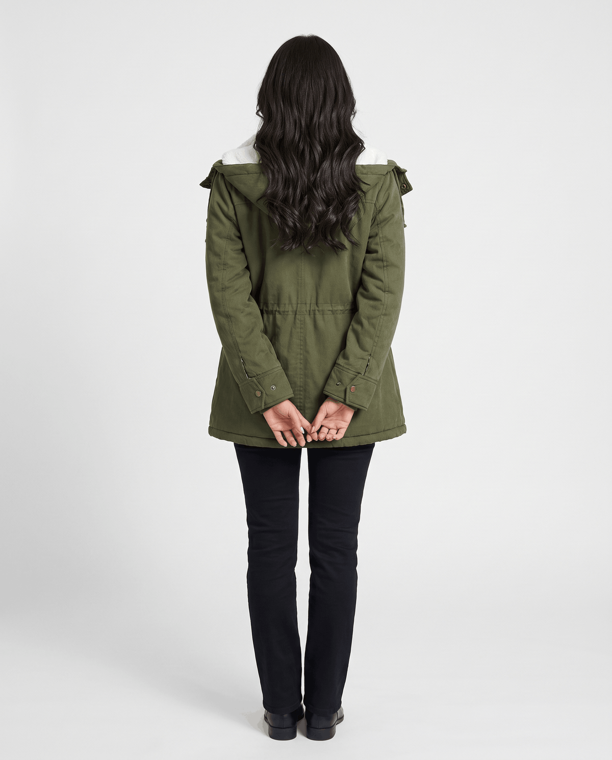 A woman wearing an olive green mid-length parka jacket with a fur-trimmed hood and drawstring waist, shown from the back