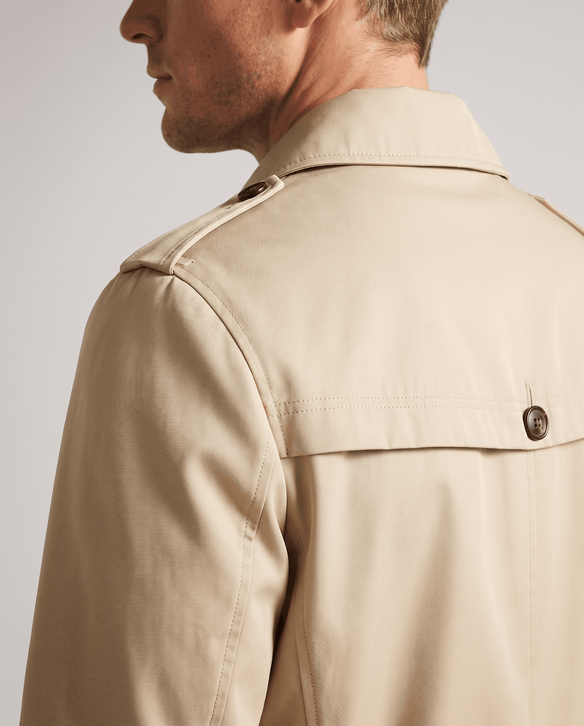 Close-up of a man wearing a beige mid-length single-breasted trench coat, showing the collar and back detail