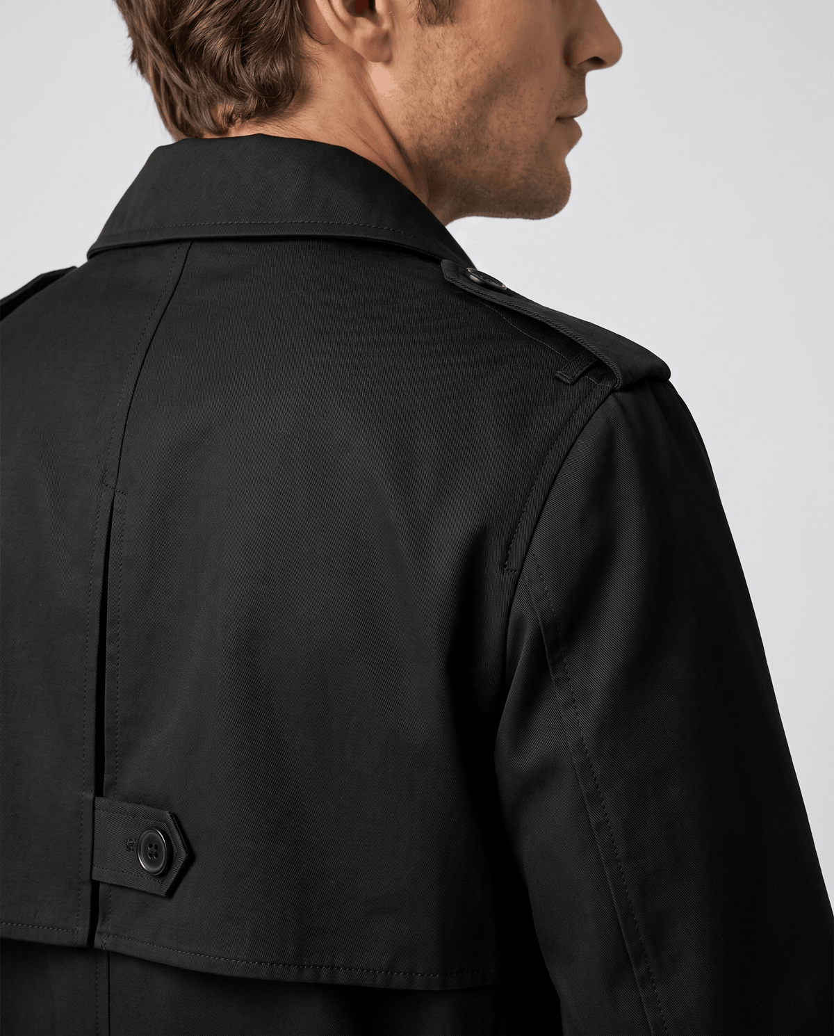 A man wearing a black mid-length single-breasted trench coat with clean lines, shown from the back with a buttoned back vent