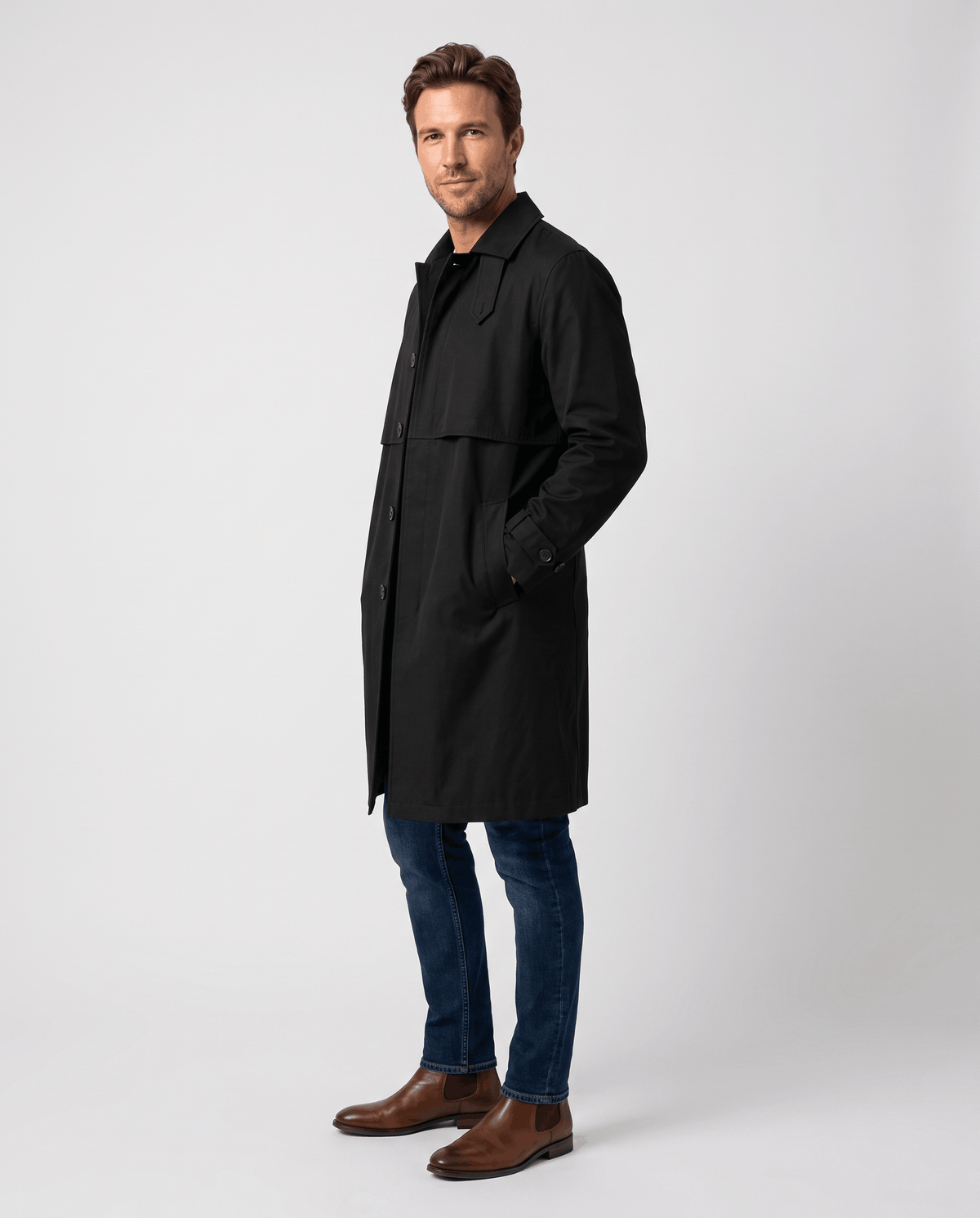 A man wearing a black mid-length single-breasted trench coat with a button front and clean lines, side view