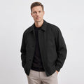 Male model wearing a black collared jacket over a black shirt and light trousers, standing with hands in pockets.