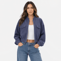Female model wearing a blue cropped utility jacket with buttoned chest pockets, a stand collar, and elastic cuffs, paired with blue jeans.