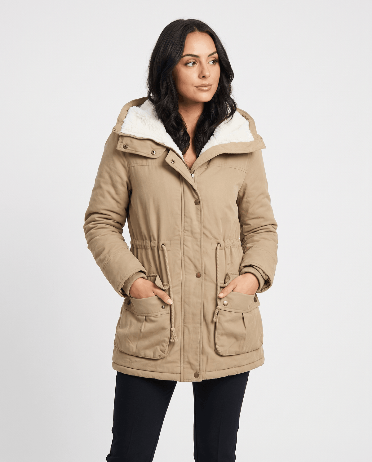 A woman wearing a beige mid-length parka jacket with plush lining, a hood, and adjustable waist, full-body view