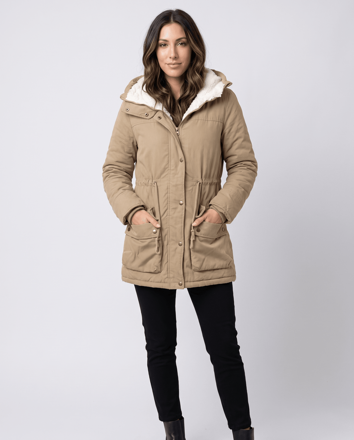 A woman wearing a beige parka jacket with plush lining, a mid-length fit, hood, and adjustable waist, adjusting the front