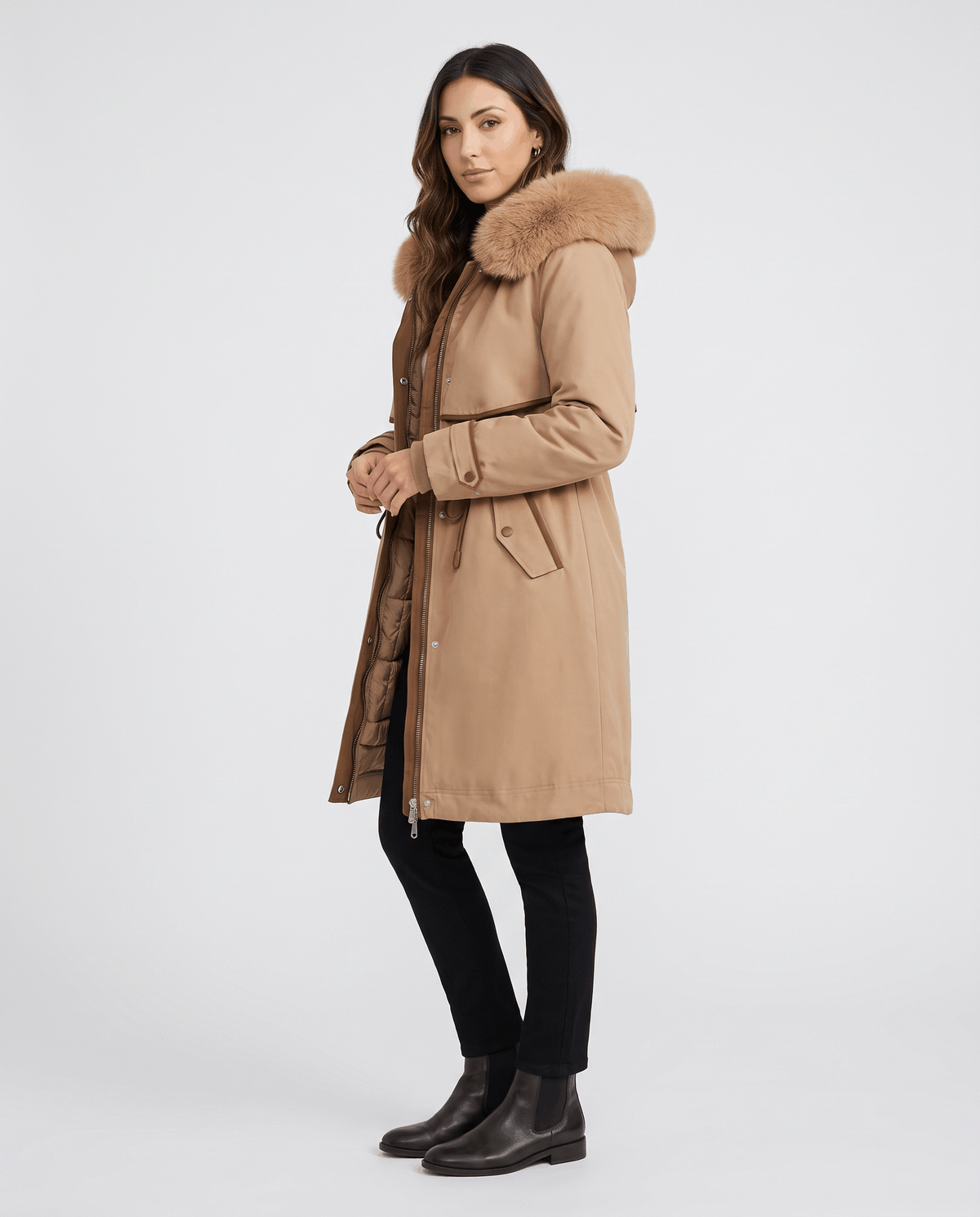 A woman wearing a beige winter parka with a plush fur-trimmed hood and adjustable waist, shown in side view