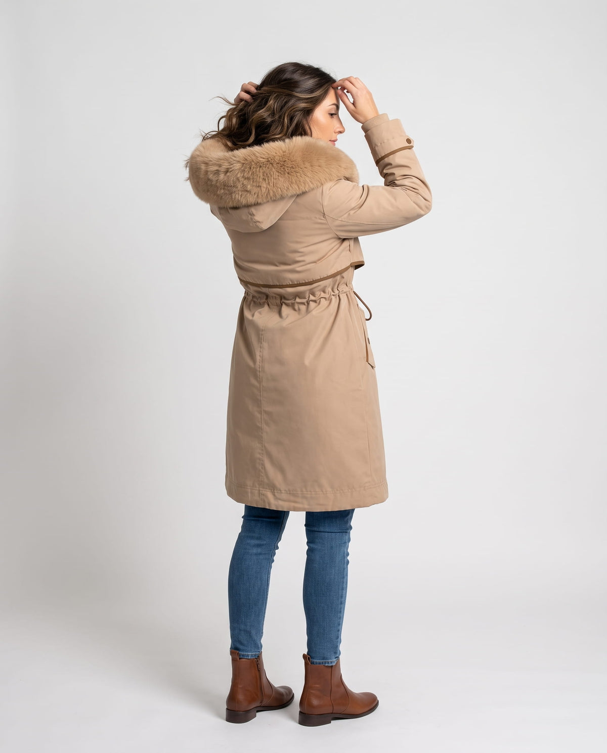 A woman wearing a beige winter parka with a plush fur-trimmed hood and adjustable waist, adjusting the hood