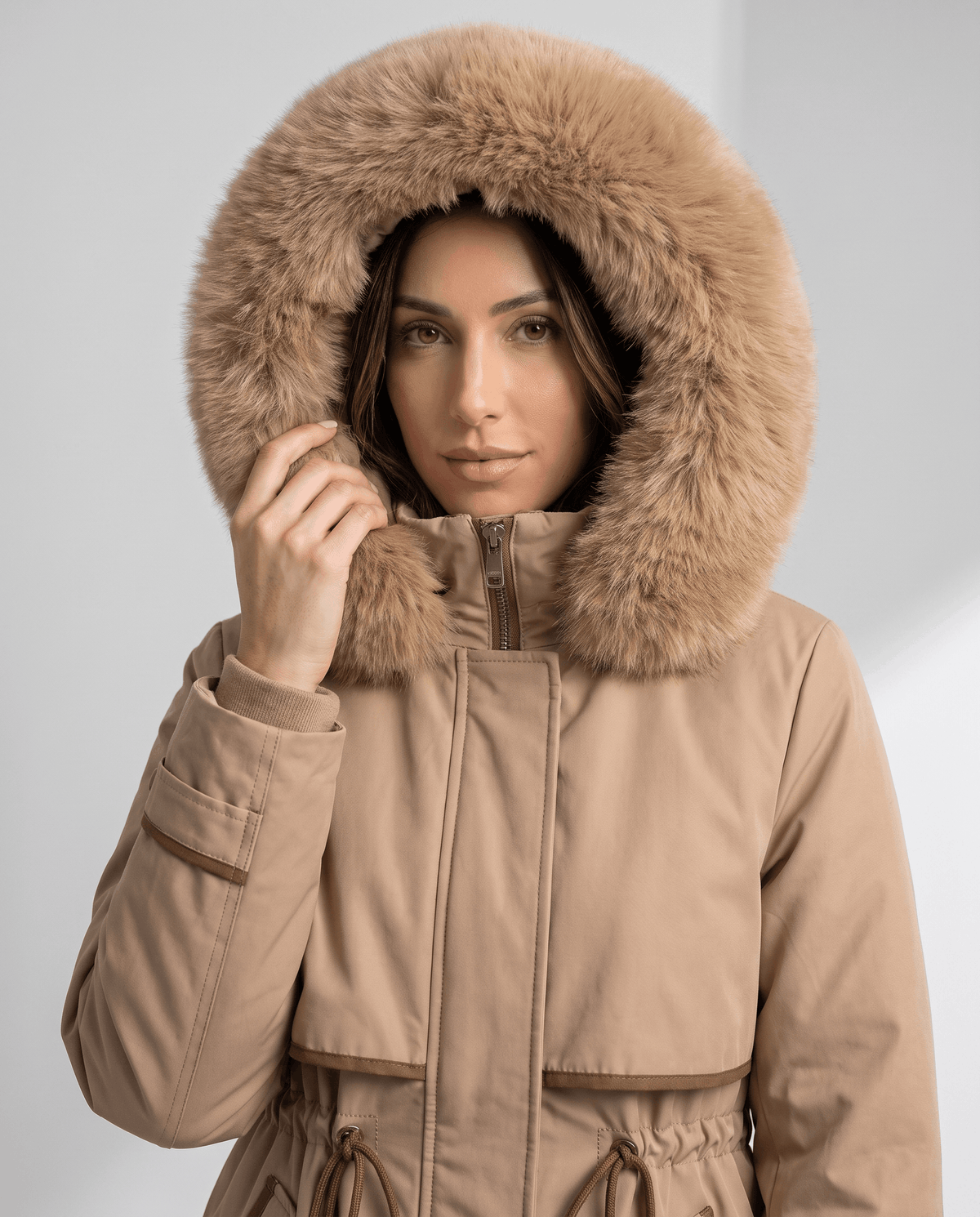 A woman wearing a beige winter parka with a large plush fur-trimmed hood and adjustable waist, front view