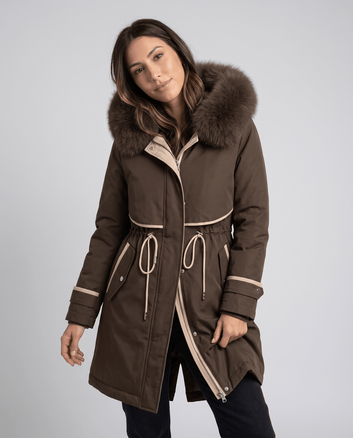 A woman wearing a brown winter parka with a plush fur-trimmed hood and adjustable waist, shown in three-quarter view