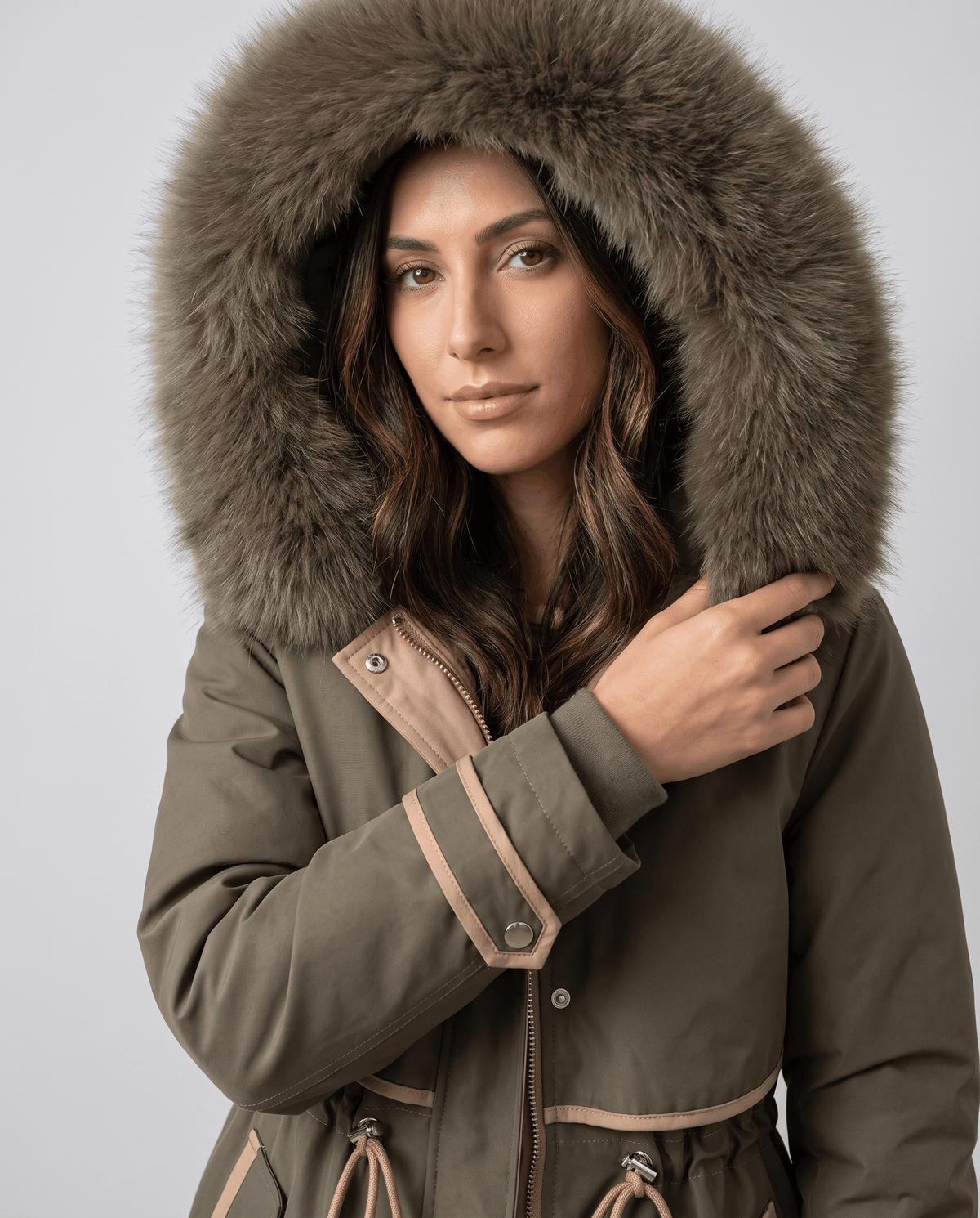 A woman wearing an olive green winter parka with a plush fur-trimmed hood, adjusting the hood and waist