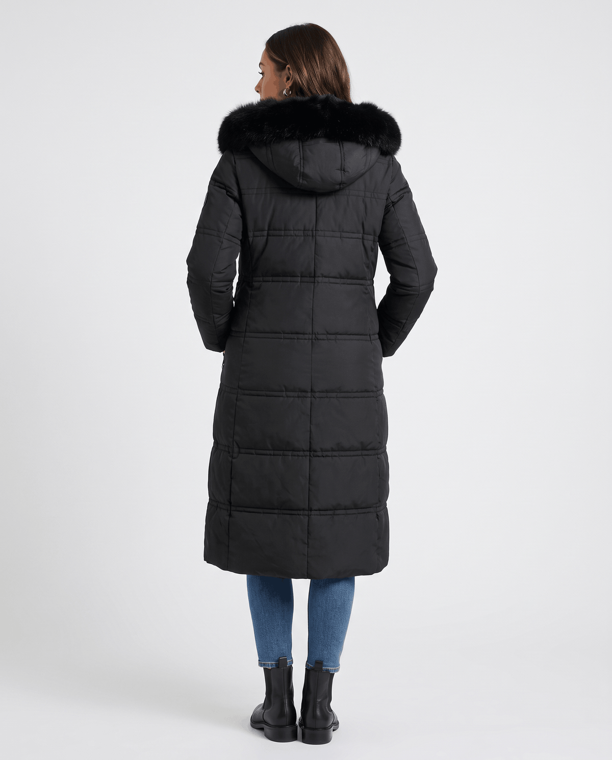 A woman wearing a black longline quilted winter parka with a plush fur-trimmed hood, shown from the back