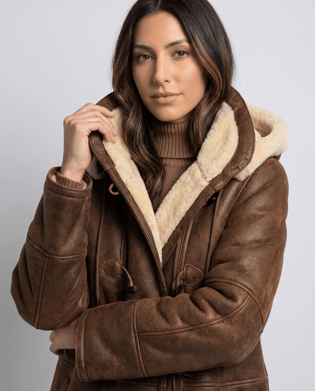 A woman wearing a dark brown shearling duffle coat with a hooded design and toggle zip closure, front view