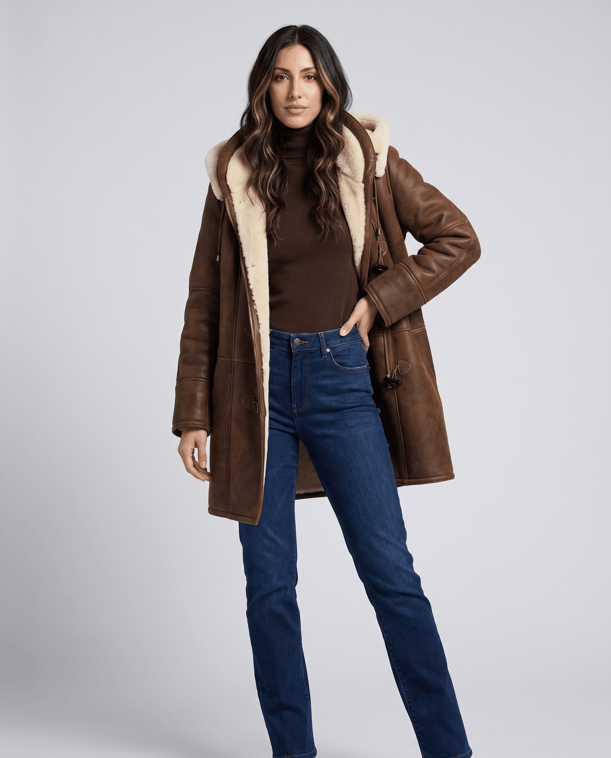 A woman wearing a dark brown shearling duffle coat with a hood and toggle zip closure, standing indoors