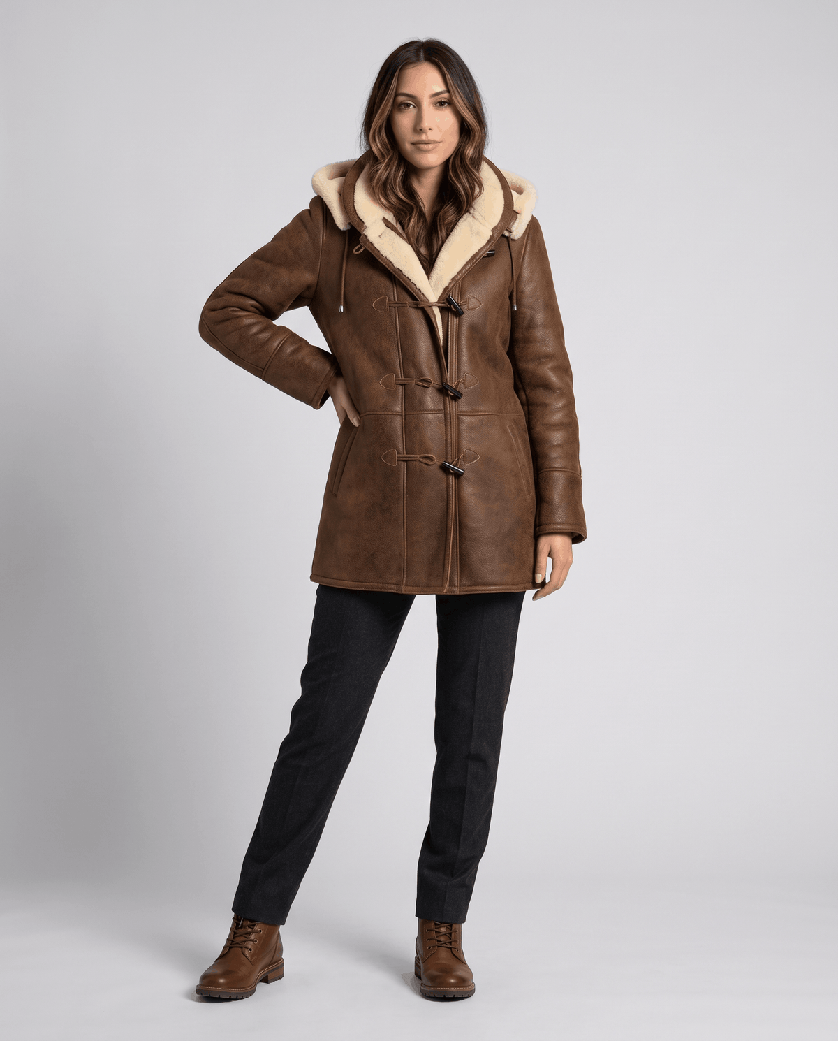 A woman wearing a dark brown shearling duffle coat with a hood and toggle zip closure, shown in full-length view