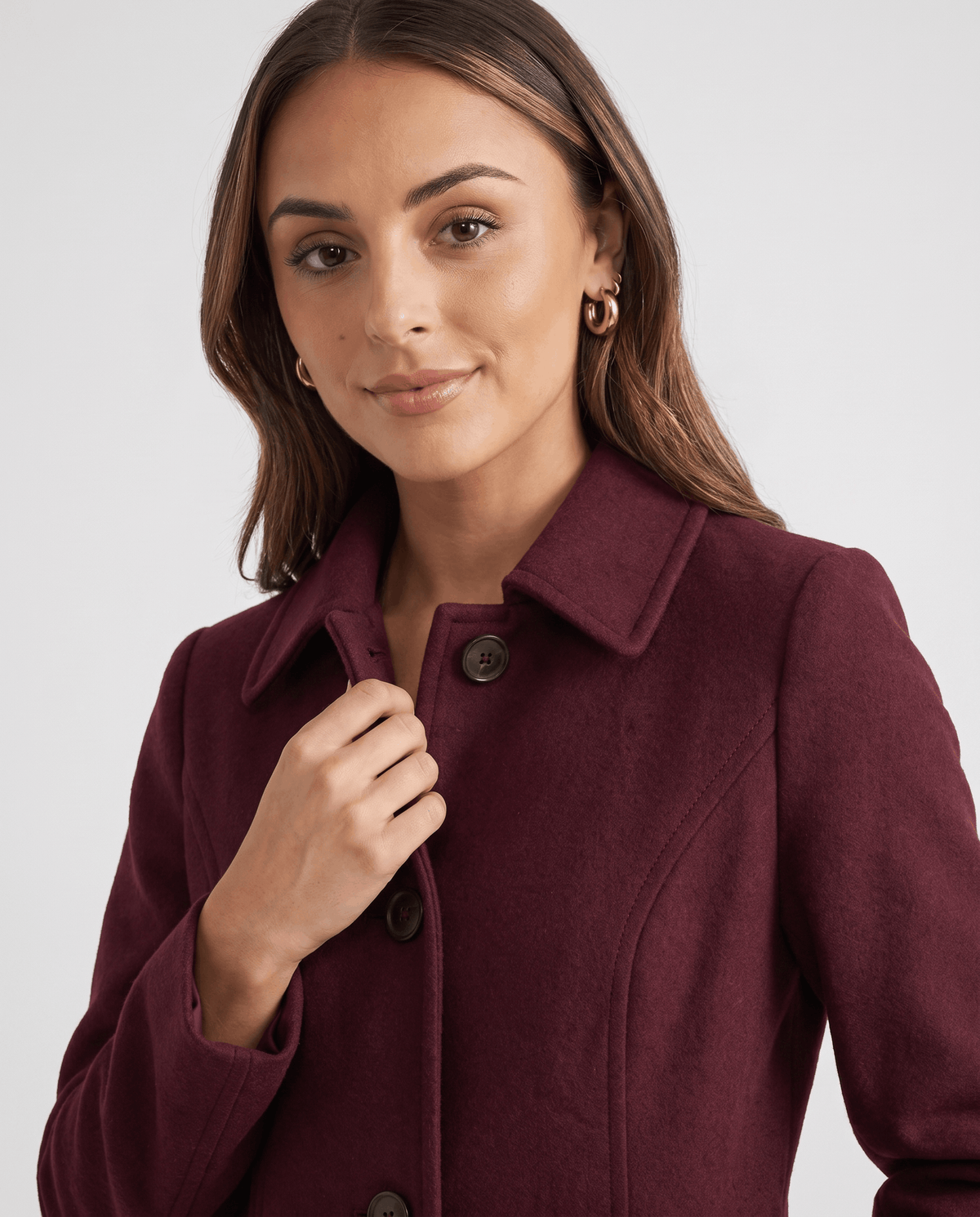 Close-up of a woman wearing a burgundy single-breasted mid-length trench coat, showing the lapel collar and button front