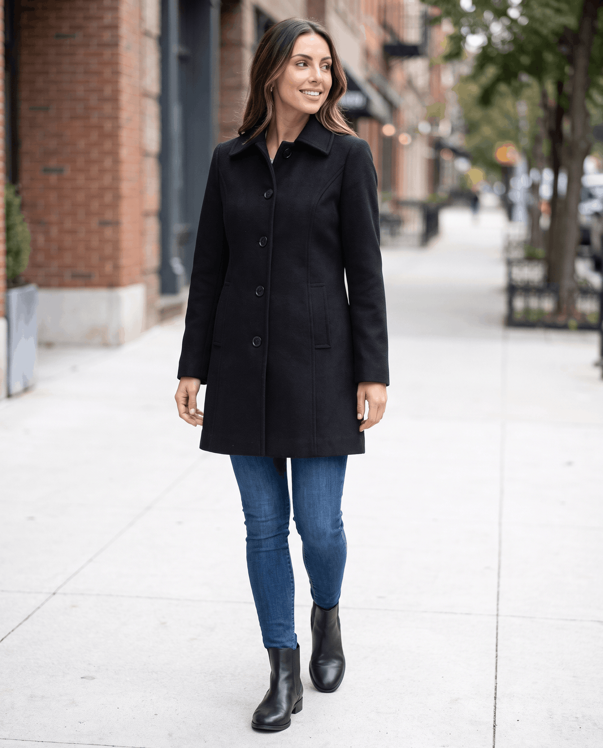 A woman wearing a black single-breasted mid-length trench coat with a button front, walking outdoors