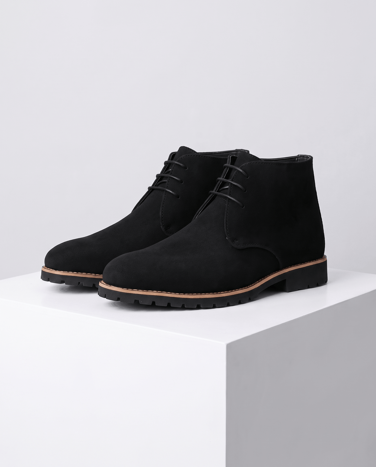 A pair of black suede ankle boots with lug soles and three-eyelet lacing, shown on a display stand