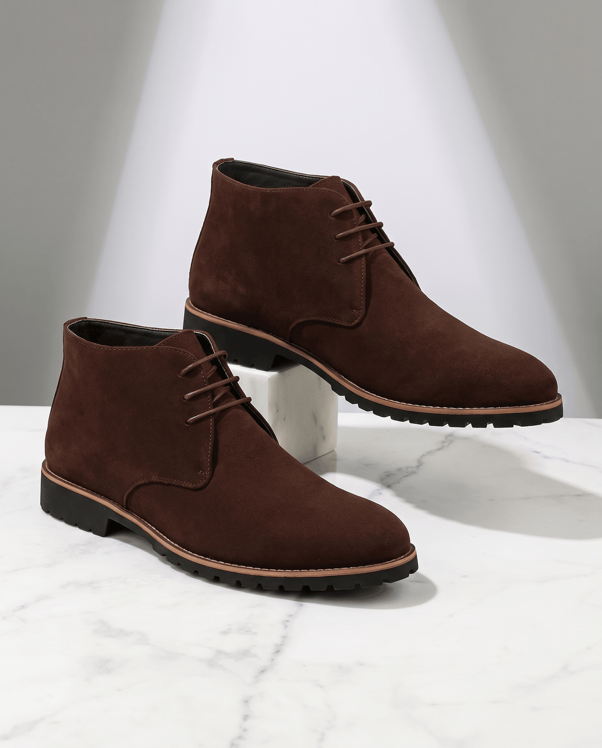 A pair of brown suede ankle boots with lug soles and three-eyelet lacing, shown on a marble surface