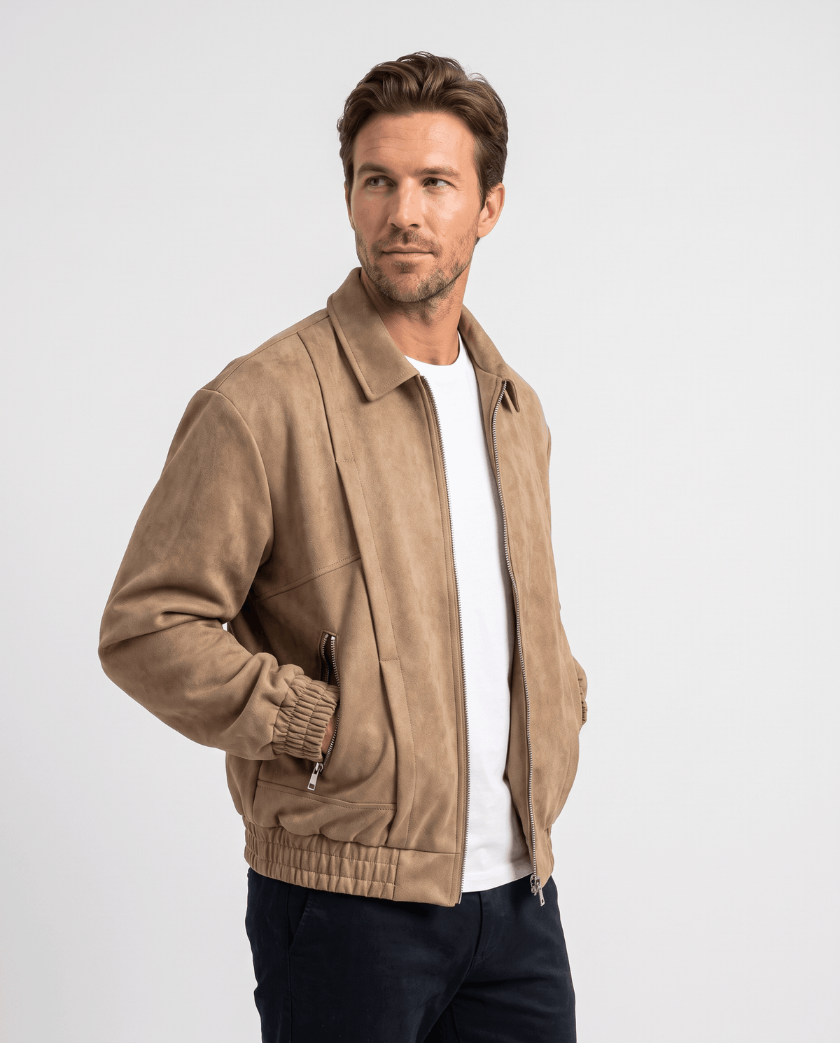 A man wearing a brown suede bomber jacket with a relaxed fit, zip pockets, and stand collar, side view