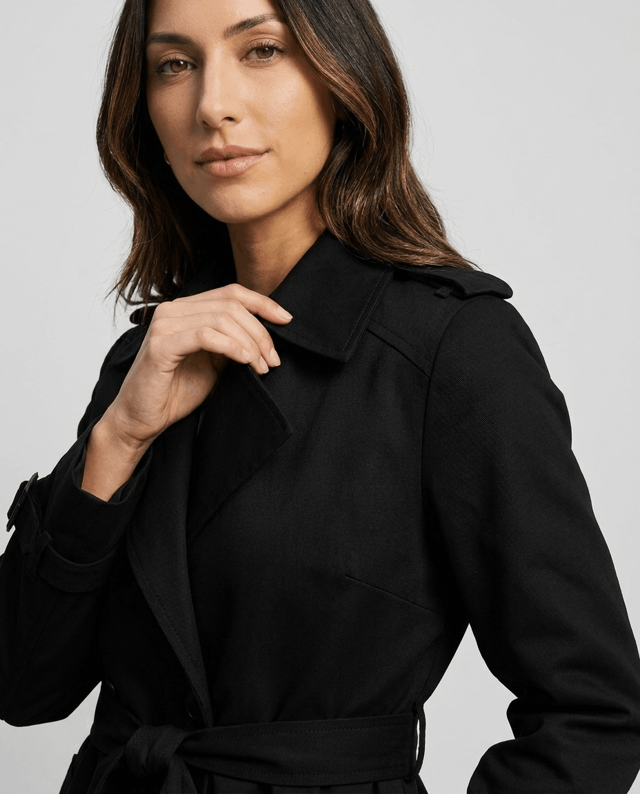 A woman wearing a black mid-length trench coat with a lapel collar and belted waist, close-up view