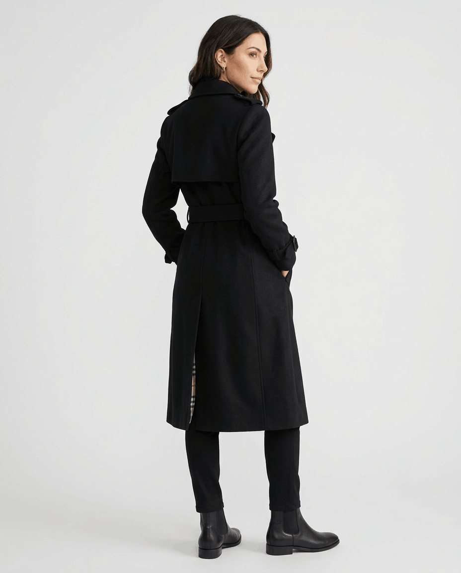 A woman wearing a black mid-length trench coat with a belted waist, shown from the side