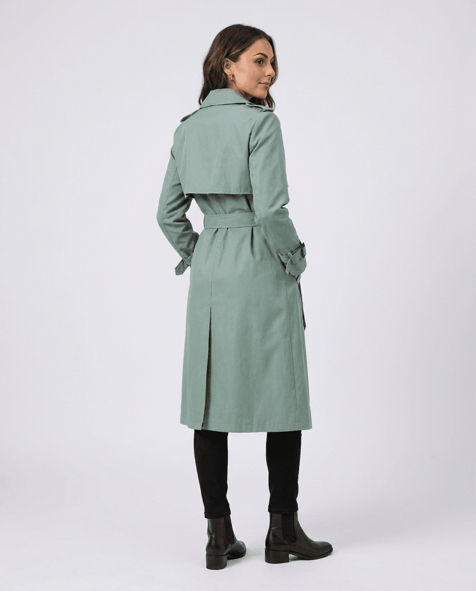 A woman wearing a sage green mid-length trench coat with a belted waist, shown from the back