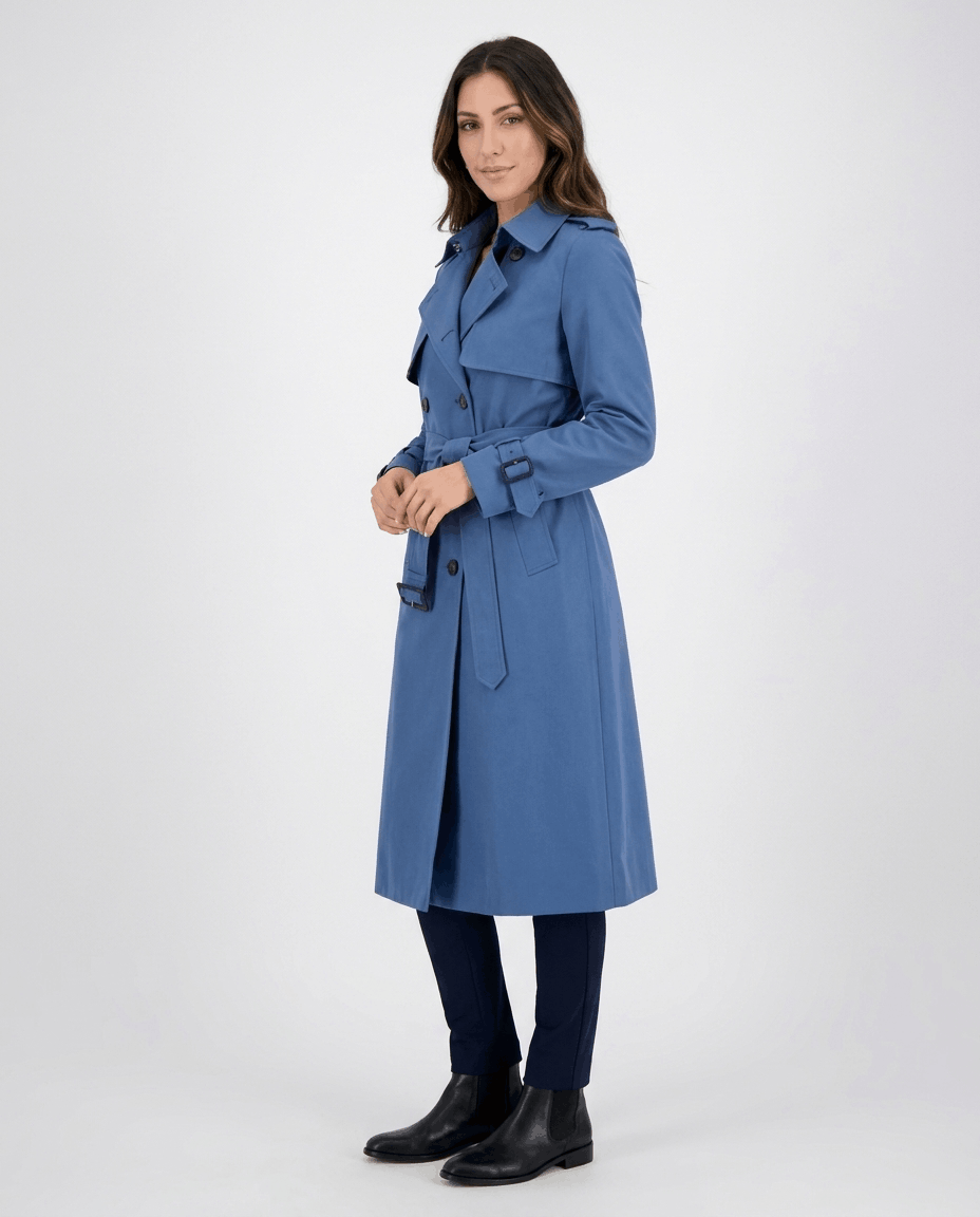 A woman wearing a blue mid-length trench coat with a belted waist, button front, and side pockets