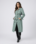 A woman wearing a sage green mid-length trench coat with a belted waist, button front, and side pockets