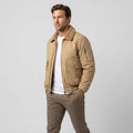 Male model wearing a beige bomber jacket with a contrasting shearling-style collar, buttoned chest pockets, and a sleeve pocket, paired with a white shirt and brown trousers.