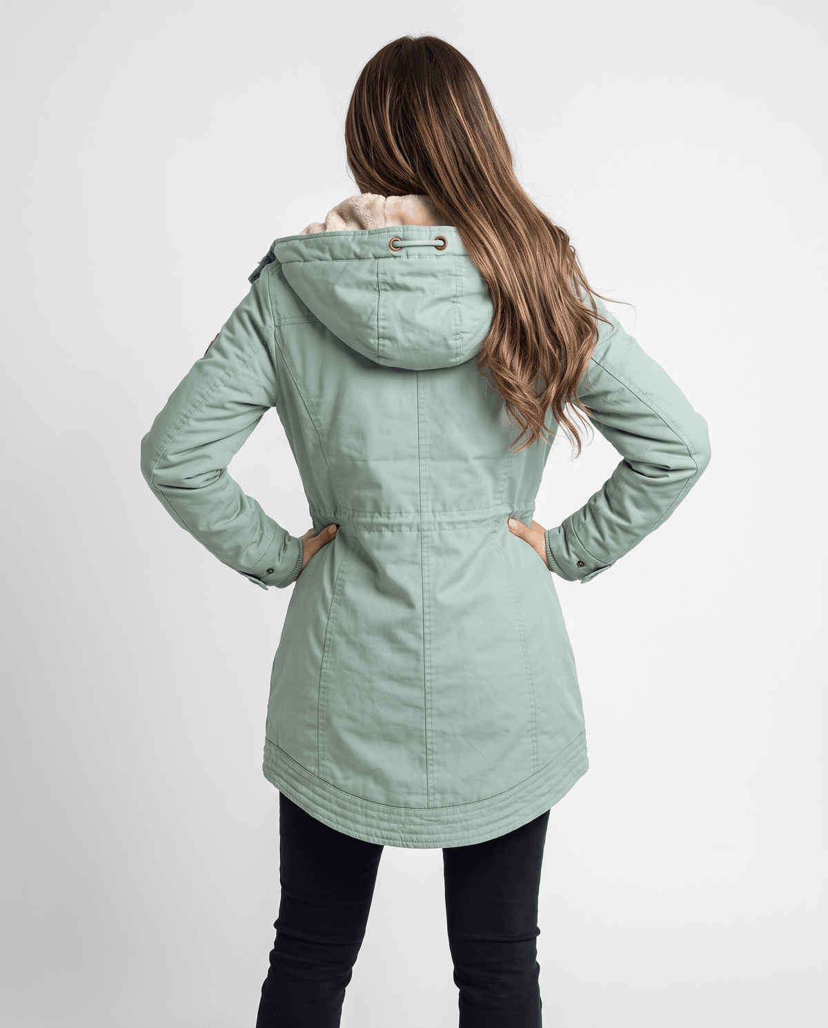 A woman wearing a light green winter parka jacket with a hood, plush lining, and drawstring waist, shown from the back