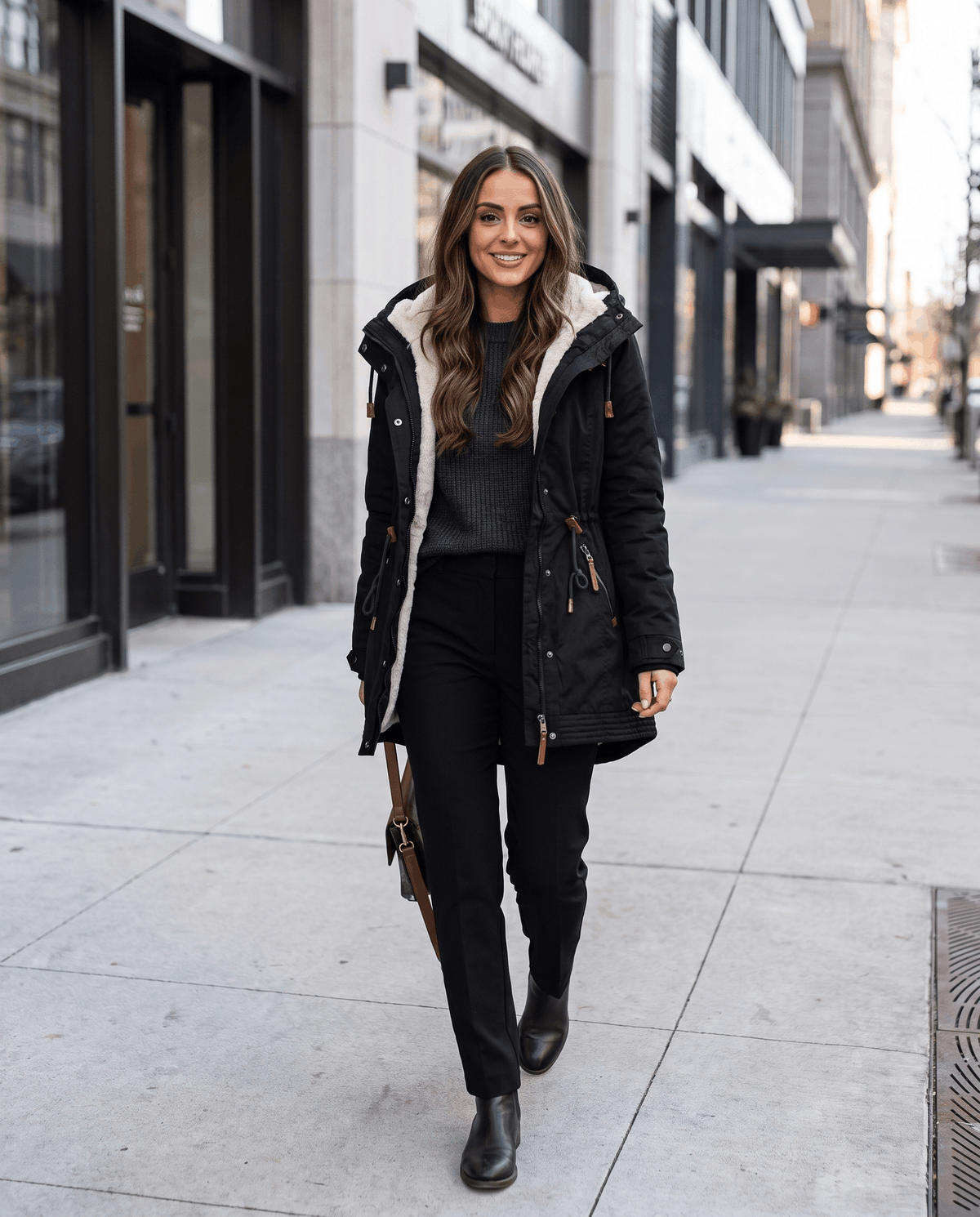 A woman wearing a black winter parka jacket with a hood, plush lining, and drawstring waist, walking on a city street
