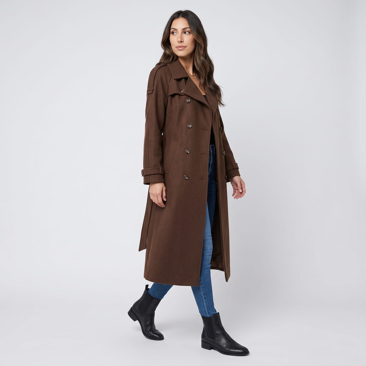 A woman wearing a brown wool trench coat with a double-breasted front and waist belt, shown in side view