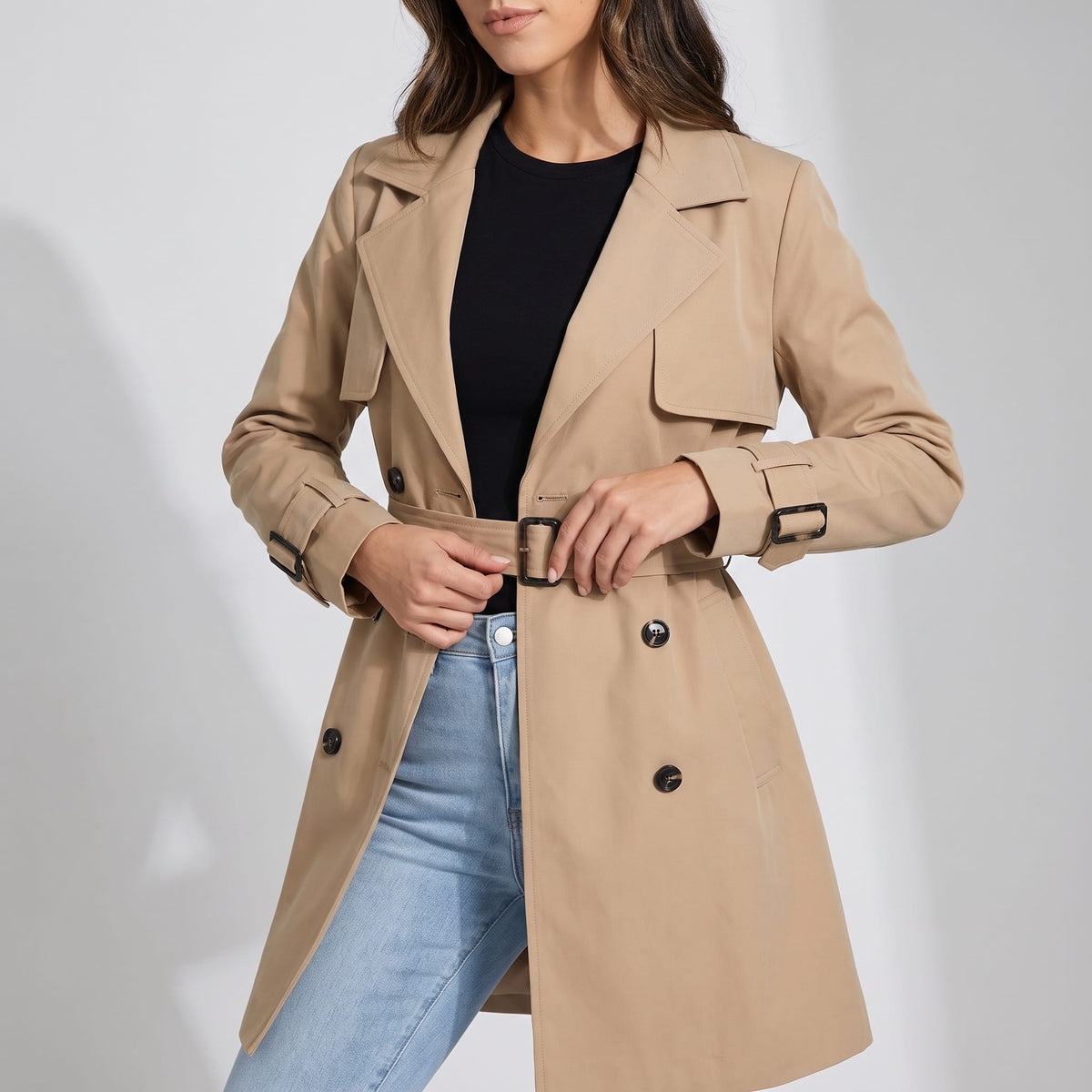 Beige coat worn open over a black top and light jeans, belt in hand