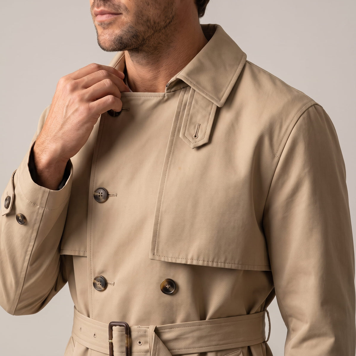 Close-up of beige trench coat buttons, tie belt and cuff strap, model touching collar
