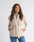 Belted puffer vest in beige with shawl collar, tied at the waist, model hands in pockets