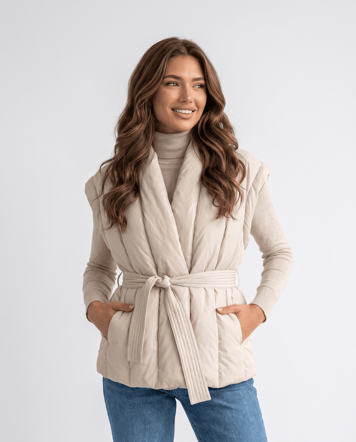Belted puffer vest in beige with shawl collar, tied at the waist, model hands in pockets