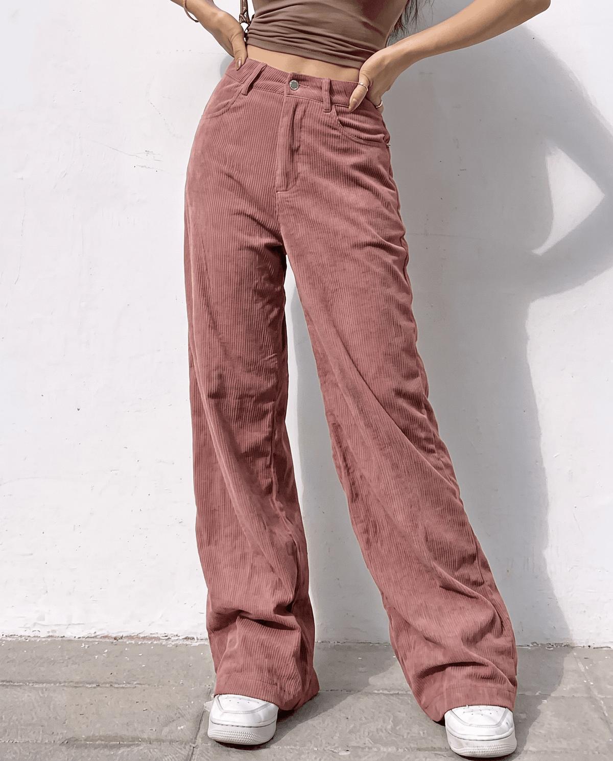 Woman wearing dusty pink high-waisted cotton corduroy wide-leg pants and white sneakers against a white wall