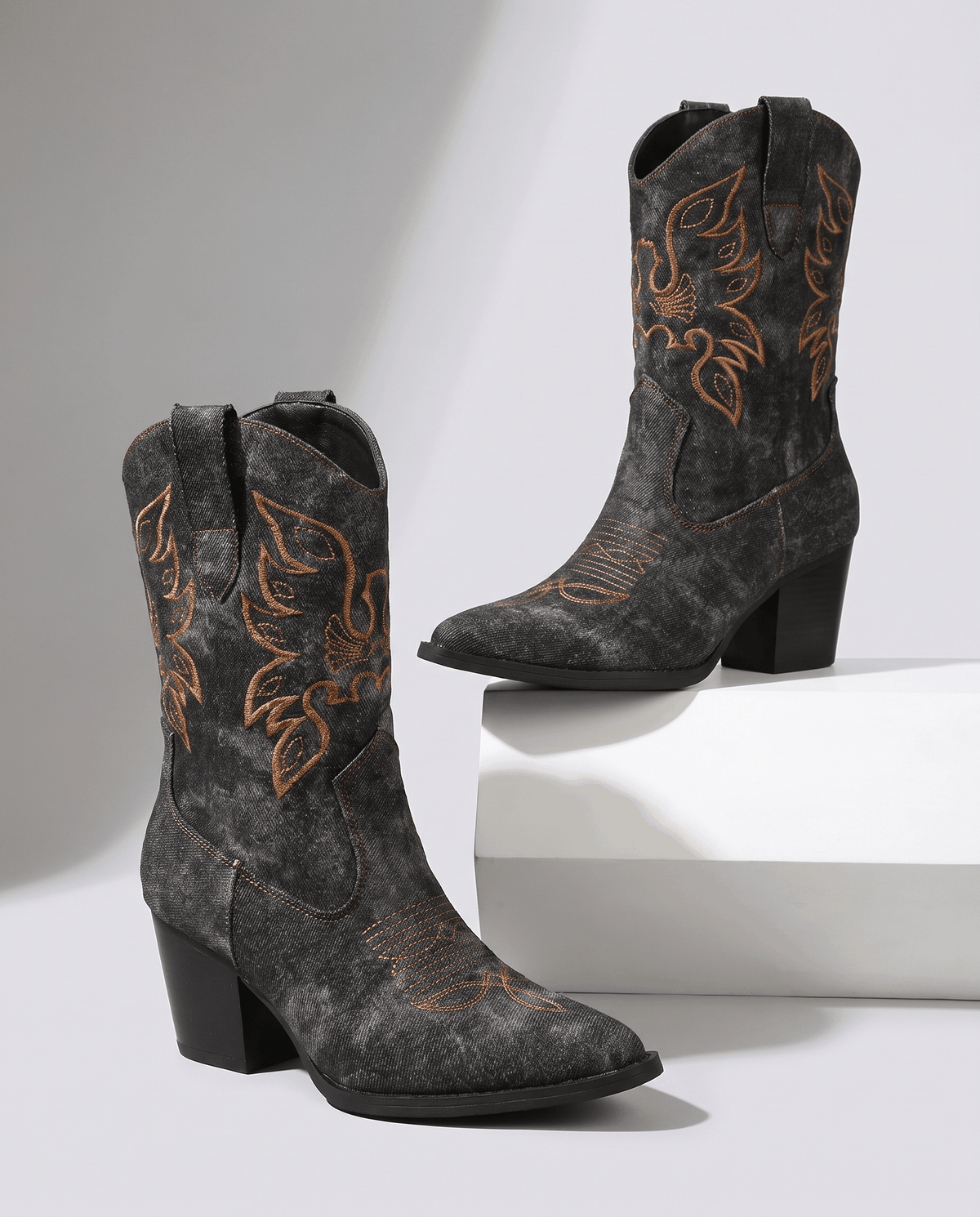 Two black denim-look boots with golden embroidery, one resting on a white block
